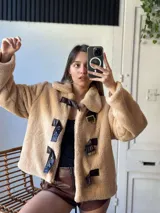 Campera corta color beige de tela tipo peluche con cuello solapa y cierre frontal con presillas de cuero sintético y hebillas metálicas.