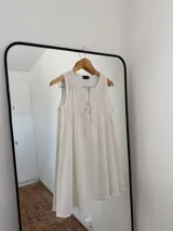 Vestido blanco sin mangas, con escote redondo y detalle de pliegues verticales en el frente, con lazos ajustables en el escote. Corte amplio y holgado.
