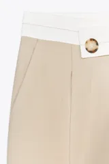 Pantalón color beige de tiro alto con cintura combinada en blanco, pinzas delanteras y pernera ancha. Cierre frontal con cremallera y botón.