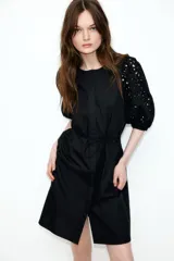 Vestido a la rodilla negro en tejido liviano de algodón con cuello redondo, botones ocultos adelante y cinturón para atar fino extraíble. Mangas al codo abullonadas en broderie con puños elásticos.