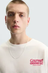 Camiseta blanca de corte holgado, cuello redondo y manga corta. Estampado frontal con la frase "Gourmet Groove LLC" en color fucsia.