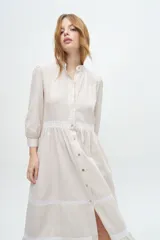 Vestido camisero midi color beige a rayas verticales, con cuello mao, mangas 3/4 abullonadas y festones de algodón en el cuerpo. Cierre frontal con botones.