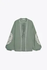 Blusa verde salvia de algodón con cuello redondo y escote pico, manga larga con encaje a contraste, puntilla y pliegues en el delantero. Cierre frontal con botones y lazada acabada en pompones.