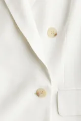 Blazer blanco de doble botonadura con solapas de muesca, hombreras y bolsillos delanteros ribeteados con solapa.