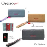Billetera color camel con textura grabada con el texto "The Lovers Oreiro Love". Incluye correa de muñeca con el texto "Oreiro Love" y un dije circular rojo.