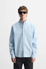 Camisa celeste de corte regular fit, confeccionada en popelín de algodón. Tiene cuello abotonado, manga larga con puños abotonados y cierre frontal con botones.