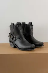 Botas cortas de cuero color marrón oscuro, con puntera cuadrada y caña baja. Presentan una correa con hebilla metálica alrededor del tobillo y un estribo desmontable.