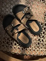 Sandalias de cuero color beige con doble tira, hebillas metálicas plateadas, tachas pequeñas y suela plana negra.