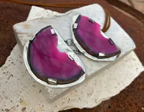 Collar con colgante de dos piedras naturales irregulares engarzadas en metal plateado y dorado, suspendidas de un cordón de cuero marrón con detalle de nudo corredizo. Las piedras tienen vetas en tonos naranja, marrón y blanco.