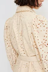 Vestido corto color beige con broderie, cuello camisero, botonadura frontal, cinturón extraíble con hebilla redonda, mangas globo largas con puños abotonados y ribetes festoneados.