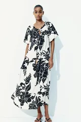 Vestido largo de corte holgado con estampado floral en blanco y negro. Tiene escote cuadrado con cordón ajustable y mangas cortas abullonadas con elástico en los puños. Presenta costura fruncida bajo la cadera.