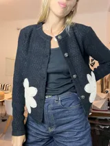 Saco de lana negro, de corte clásico y abotonado, con cuello redondo. Presenta apliques de flores de crochet blancas en la parte delantera inferior. Los botones son redondos y plateados con un diseño grabado.