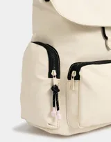 Mochila color beige con detalles en negro. Tiene solapa con tira de ajuste y broche, correas regulables, manija superior, bolsillo frontal con cierre y puller de cordón y goma, bolsillos laterales con cierres y pullers de cordón y goma, y tag de goma.