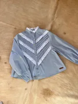 Camisa celeste con cuello alto con volados y cierre frontal con botones. Presenta detalles de encaje blanco en forma de V en el frente y mangas largas con puños abotonados.