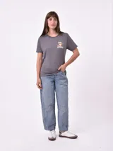 Remera blanca de algodón con cuello redondo y manga corta. Presenta un pequeño bordado en el pecho izquierdo con una figura que parece ser una persona acostada en una tabla de surf o flotador.