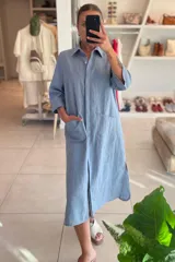 Vestido camisero color celeste de lino, con cuello clásico, cierre frontal con botones y mangas 3/4. Tiene dos bolsillos de parche en el pecho y corte irregular en el ruedo.