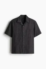 Camisa de manga corta color gris oscuro con rayas verticales, cuello resort y bolsillo de parche en el pecho. Corte recto y cierre con botones.