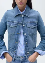 Chaqueta de jean celeste con cuello camisero, cierre frontal con botones metálicos, dos bolsillos de parche con solapa en el pecho y mangas largas con puños abotonados.