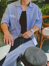 Camisa de manga corta con corte híbrido boxy fit/oversize, confeccionada en algodón. Disponible en tres diseños: a cuadros marrones, a rayas blancas y negras, y a rayas burdeos y blancas. Presenta bordados de diseño propio de la marca.