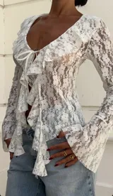 Blusa blanca de encaje floral transparente, con escote en V, cierre frontal con lazos y volantes en el escote y en el frente.