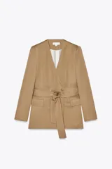 Blazer color beige con escote en pico sin solapa, manga larga con hombreras, bolsillos delanteros con solapa, cierre frontal cruzado con botones forrados a tono y cinturón del mismo tejido.