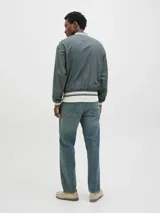 Campera bomber casual color gris verdoso, con cuello tipo baseball y puños de canalé a rayas blancas. Cierre con botones a presión y bolsillos frontales. Confeccionada en tejido liviano y transpirable con hombros caídos.