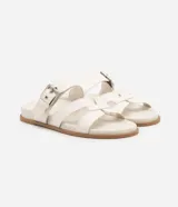 Sandalias planas de cuero sintético color blanco, con puntera redonda y dos tiras, una con hebilla metálica y otra en forma de H.