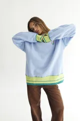 Buzo celeste con cuello polo y puños en rib color verde lima. Tiene un parche con el logo de la marca en el pecho y rayas horizontales en el ruedo.