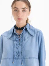 Camisa de denim azul claro, manga larga, con cuello clásico y escote ajustable mediante cordón que pasa por ojalillos metálicos.