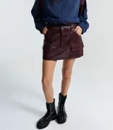 Short pollera corto color verde militar, confeccionado en poliuretano (PU) que imita el cuero. Presenta bolsillos delanteros grandes con solapas, pasadores en la cintura y cierre de cremallera.