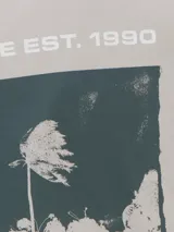 Musculosa color gris claro, de corte holgado, cuello redondo y sisas amplias. Estampado trasero con palmeras y texto "Visual Medium".