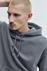 Canguro de color gris oscuro con capucha y cordón ajustable, de corte holgado y hombros caídos. Cuenta con un bolsillo frontal tipo canguro y terminaciones acanaladas en puños y bajo.