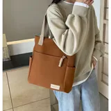 Cartera tipo tote de lona en color beige con correas de cinta marrón. Presenta un compartimento principal con cierre, un bolsillo frontal abierto y bolsillos laterales para mayor organización.