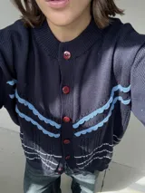 Saco de punto calado color beige claro, con cuello redondo y cierre frontal de botones azules. Presenta detalles decorativos en crochet verde y negro en el frente y puños.