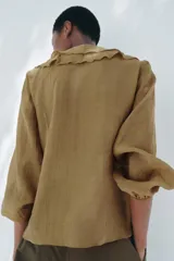 Camisa color verde oliva de tela traslúcida con escote en V, mangas largas abullonadas con puños elásticos y volantes en el frente.