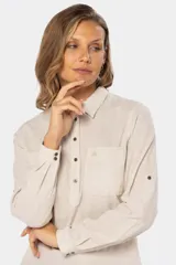 Camisa color beige de lino, de manga larga con botones metálicos personalizados y presilla en la manga. Tiene cuello clásico, cierre frontal parcial con botones y bolsillo de parche en el pecho.