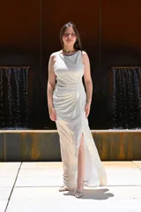 Vestido largo de fiesta color gris claro metalizado, con escote redondo, sin mangas y abertura lateral en la falda. Presenta un drapeado lateral que estiliza la figura.