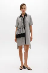 Vestido camisero a rayas verticales en tonos grisáceos, con cuello camisero y cartera de botones negros. Presenta volados frontales y laterales, mangas cortas y bajo asimétrico.