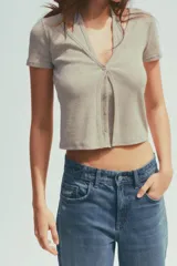 Camiseta color beige de tejido de punto, con cuello halter y escote pico. Tiene cierre frontal con botones y mangas cortas.