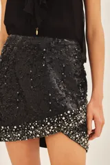 Falda corta negra de tiro medio, confeccionada en viscosa crepe, con bordado de lentejuelas y piedras en tonos plateados y blancos, concentrados principalmente en el ruedo.