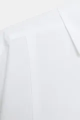 Camisa blanca de popelín con cuello solapa, manga larga y cierre frontal con botones ocultos por solapa.