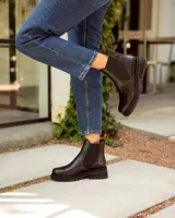 Bota tipo Chelsea de cuero negro con paneles elásticos laterales. Presenta una suela track de goma y un tirador de cuero marrón en la parte posterior.