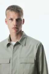 Sobrecamisa verde oliva de corte recto, con cuello inglés, cierre frontal con botones a presión y dos bolsillos de parche con solapa en el pecho.
