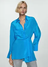 Camisa celeste de tejido satinado con cierre frontal asimétrico de botones, manga larga con puños abotonados, diseño ajustable con cuerda en la parte posterior y cuello camisero.