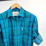 Camisa de manga larga a cuadros en tonos azul, celeste y negro. Tiene cuello clásico, cierre con botones y un pequeño logo bordado en el pecho. Las mangas pueden usarse arremangadas.