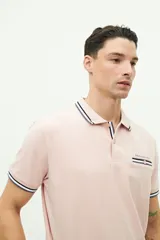 Chomba de piqué color rosa claro, de corte clásico, con cuello y mangas con ribete a rayas en blanco, gris y negro. Tiene tapeta de dos botones y logo bordado en el pecho.
