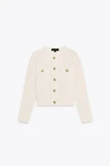 Chaqueta color crema de tejido de punto con cuello redondo y manga larga. Tiene bolsillos delanteros de plastrón y cierre frontal con botones dorados.