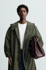 Parka verde militar de tejido técnico ligero, corte oversize, cuello alto con capucha ajustable, manga larga con puños elásticos, cintura ajustable con cordón interno, bolsillos con solapa, bajo asimétrico con abertura trasera ajustable y cierre frontal con cremallera oculta y botones a presión.