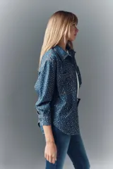 Camisa de denim azul con apliques metálicos plateados, de calce amplio, cuello camisero, cierre frontal con botones a presión, mangas largas con puños abotonados y dos bolsillos con solapa abotonada en el pecho.