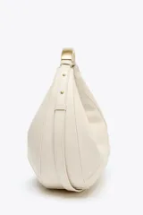 Bolso ovalado color crudo con asa de hombro metálica dorada. Tiene pespuntes en contraste, bolsillo interior con cremallera y cierre principal con cremallera y tirador.